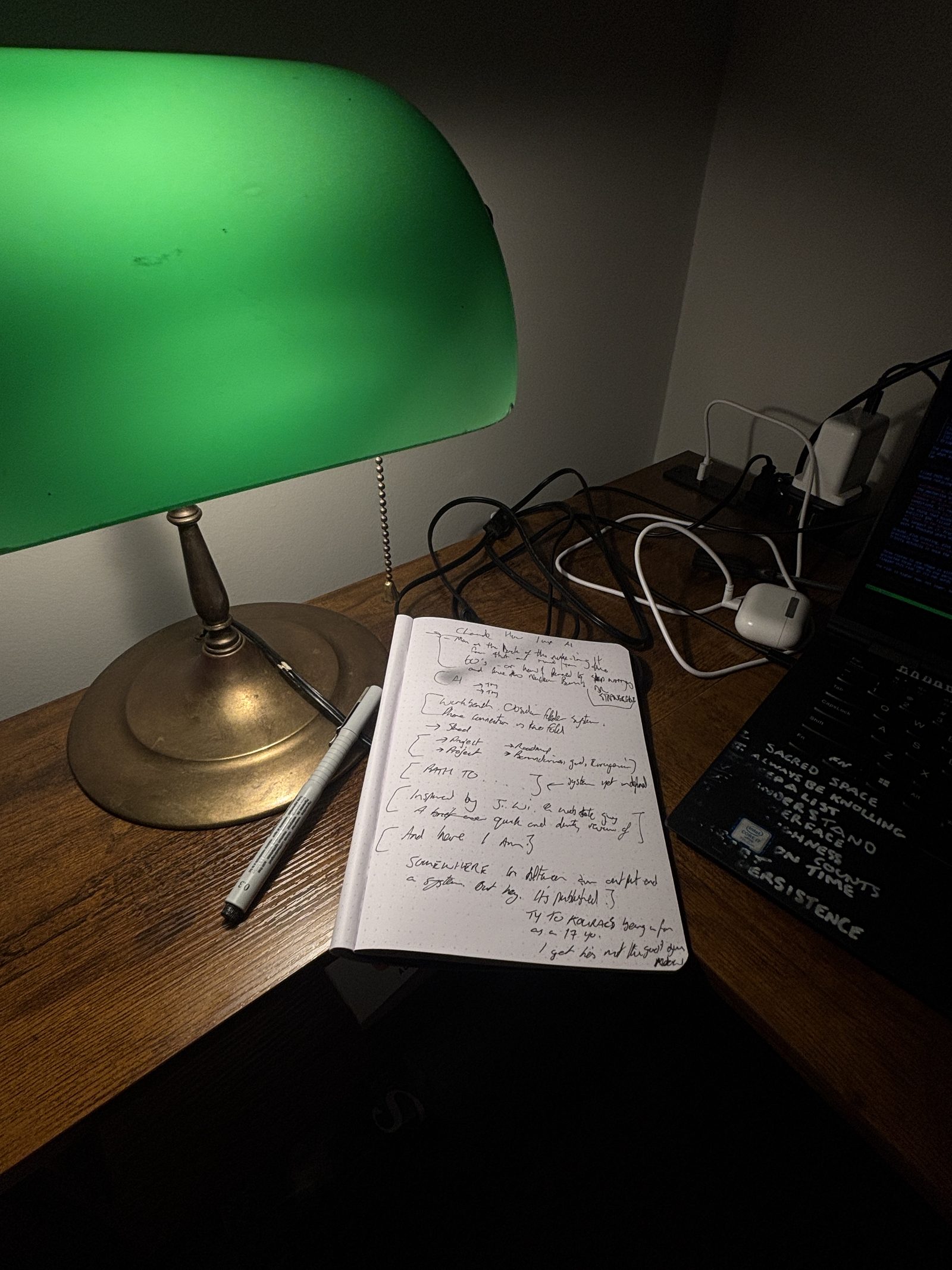 A dotted notebook open on a desk under a green banker’s lamp, covered in handwritten brackets and short phrases. A ThinkPad with words stencilled on its lid sits to the right.